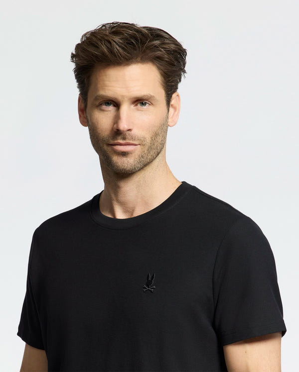 A brown-haired, stubbled man wears the black Psycho Bunny MENS TAYLOR PIQUE TEE (B6U579F200) with an embroidered chest Bunny logo against a light backdrop, gazing directly at the camera.