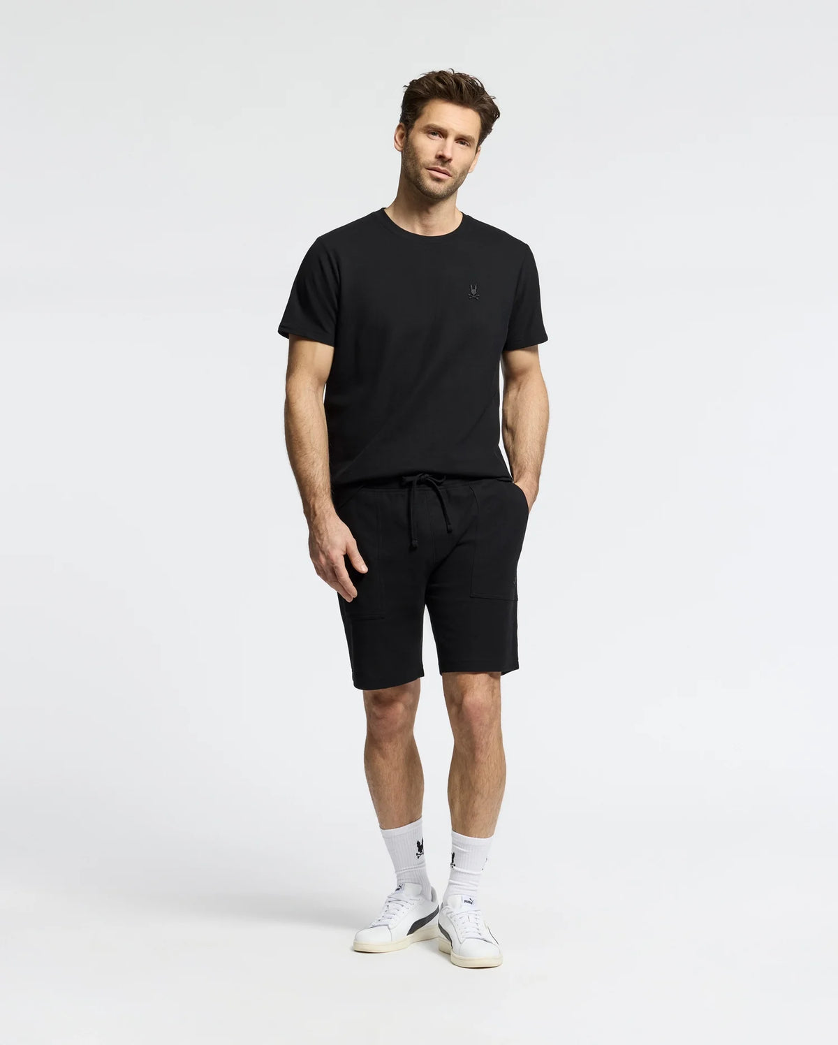 Against a white backdrop, a man wears the MENS TAYLOR PIQUE TEE by Psycho Bunny in black Pima cotton, paired with black shorts. He has his hands in his pockets and complements the look with white socks, sneakers, and a subtle embroidered Bunny on the chest for elegance.