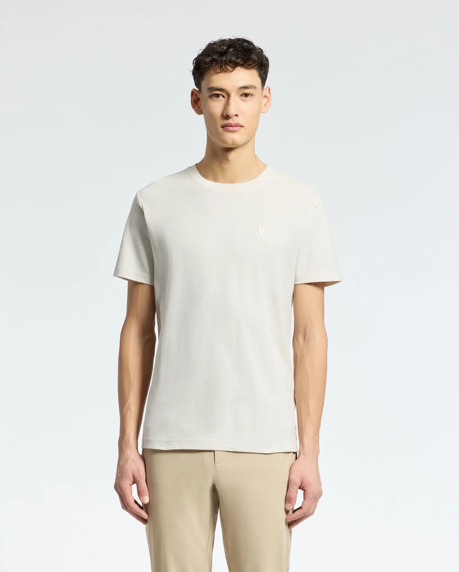 Amidst a light grey background, an individual with short curly dark hair dons a Psycho Bunny MENS TAYLOR PIQUE TEE in light beige. The regularly fit tee is made from lightweight Pima cotton and paired with beige pants, while facing forward.