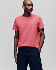 A man wears the Psycho Bunny MENS TAYLOR PIQUE TEE - B6U579F200, a short-sleeved coral pink Pima cotton shirt with an embroidered chest logo, paired with black cargo pants, standing against a plain light gray background.