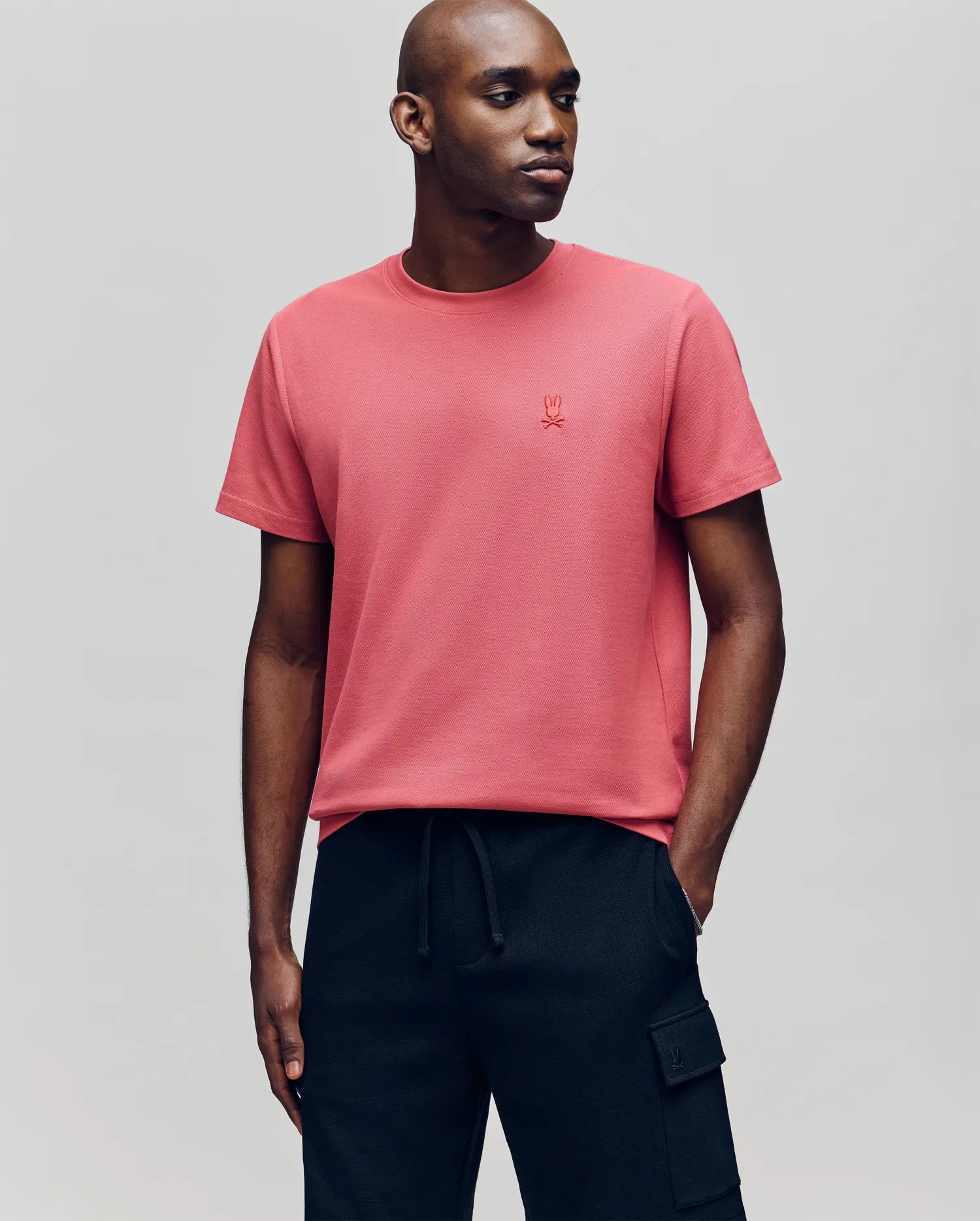 A man wears the Psycho Bunny MENS TAYLOR PIQUE TEE - B6U579F200, a short-sleeved coral pink Pima cotton shirt with an embroidered chest logo, paired with black cargo pants, standing against a plain light gray background.