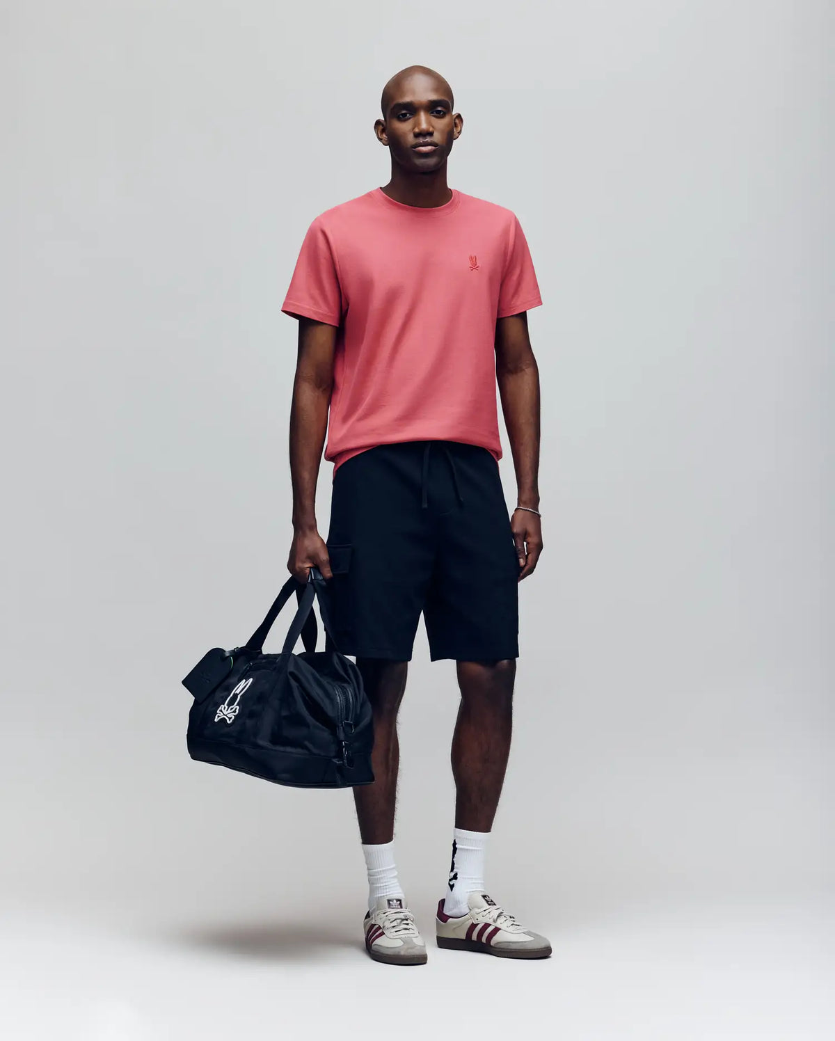 A man stands against a plain background wearing the Psycho Bunny MENS TAYLOR PIQUE TEE - B6U579F200 in pink, paired with black shorts and white sneakers. Holding a black duffel, he looks ahead with a neutral expression, showcasing modern style.