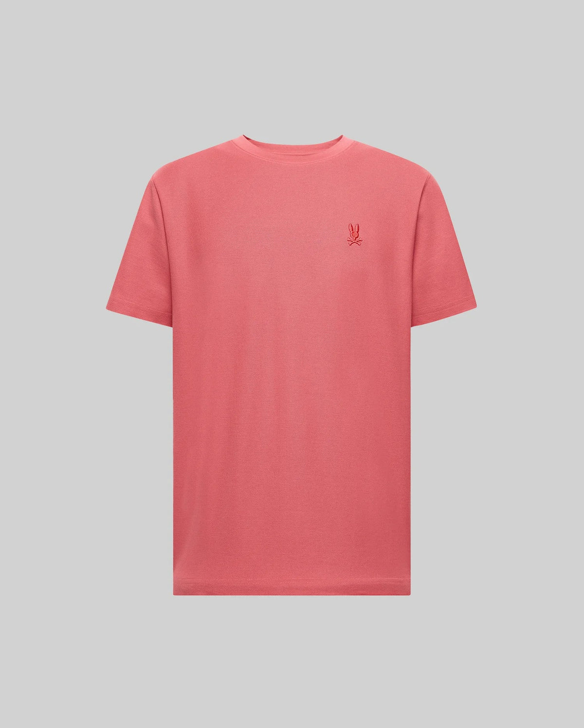 The Psycho Bunny MENS TAYLOR PIQUE TEE - B6U579F200 is a coral pink, short-sleeve Pima cotton t-shirt with a small embroidered logo on the left chest, shown against a light gray background.