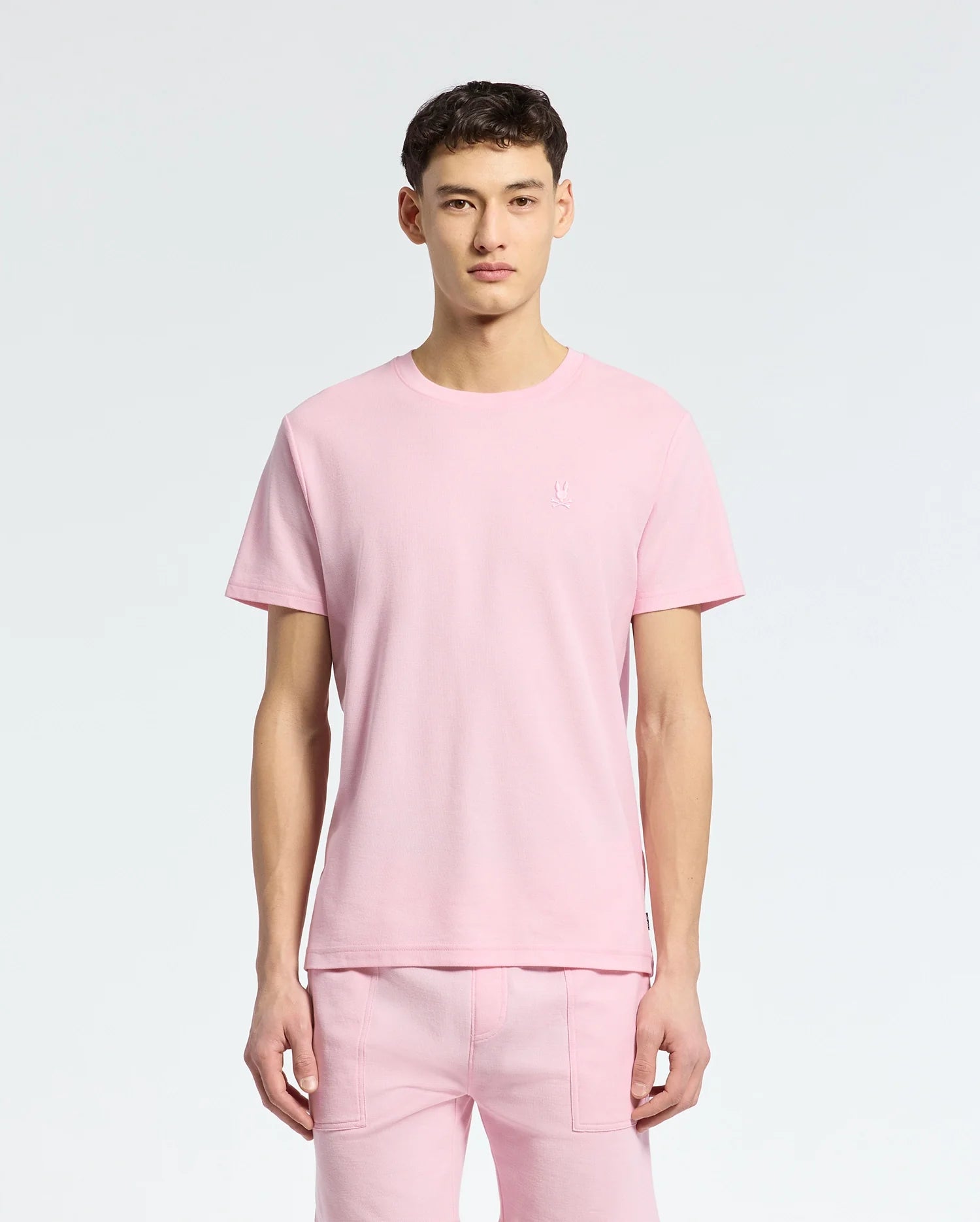 A young man faces forward in a Psycho Bunny MENS TAYLOR PIQUE TEE - B6U579F200, a light pink short-sleeved piqué tee with an embroidered chest Bunny, paired with matching pink pants against a plain light background.