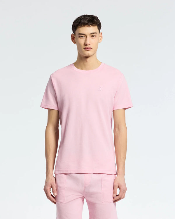 A young man faces forward in a Psycho Bunny MENS TAYLOR PIQUE TEE - B6U579F200, a light pink short-sleeved piqué tee with an embroidered chest Bunny, paired with matching pink pants against a plain light background.