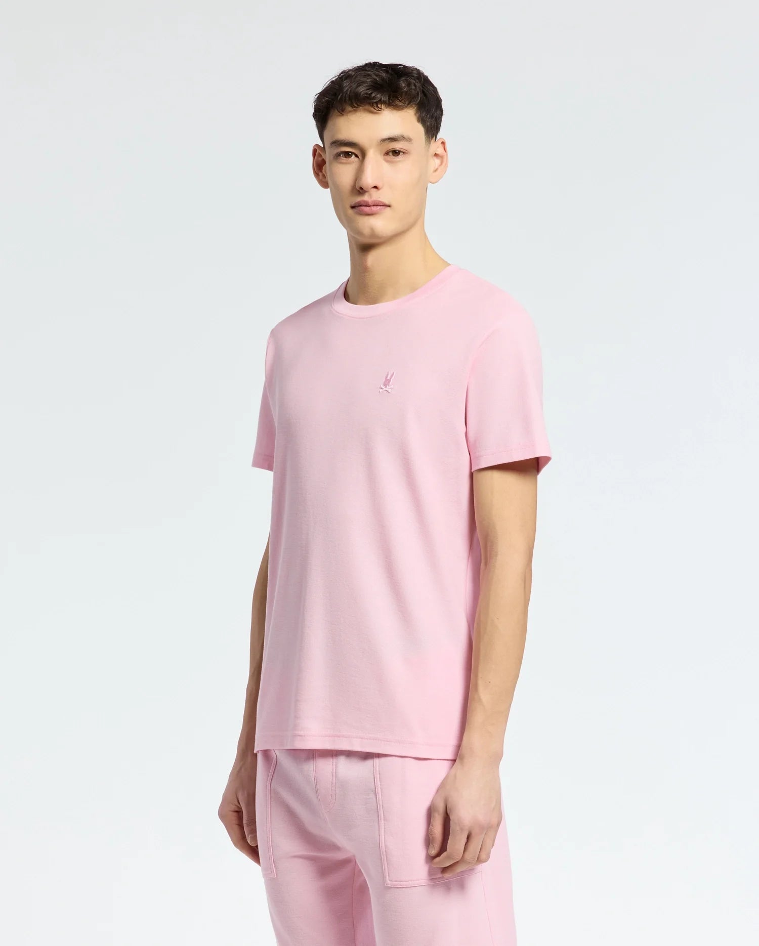 A young man faces the camera with a neutral expression, wearing the Psycho Bunny MENS TAYLOR PIQUE TEE (B6U579F200) in light pink piqué fabric, paired with matching pink pants against a plain light background.