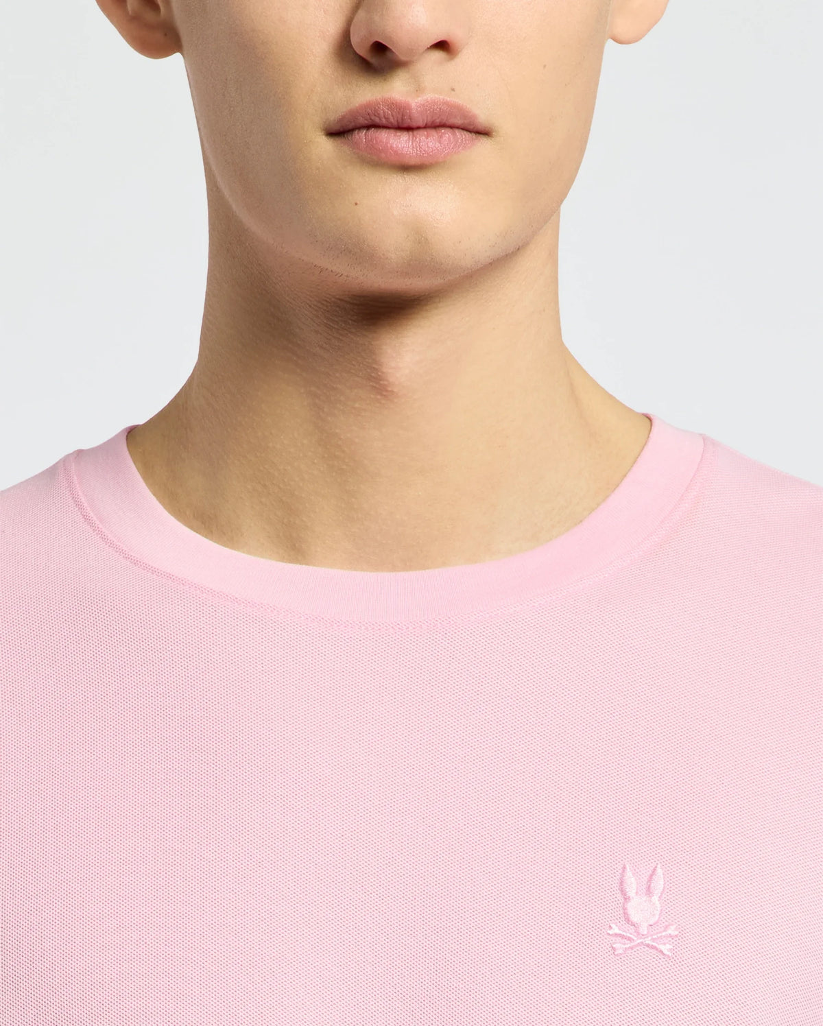 A person wears the Psycho Bunny MENS TAYLOR PIQUE TEE (B6U579F200), a light pink crew neck made from lightweight piqué, featuring a small white embroidered Bunny and crossed bones logo on the left chest. Only their lower face, neck, and upper torso are shown.