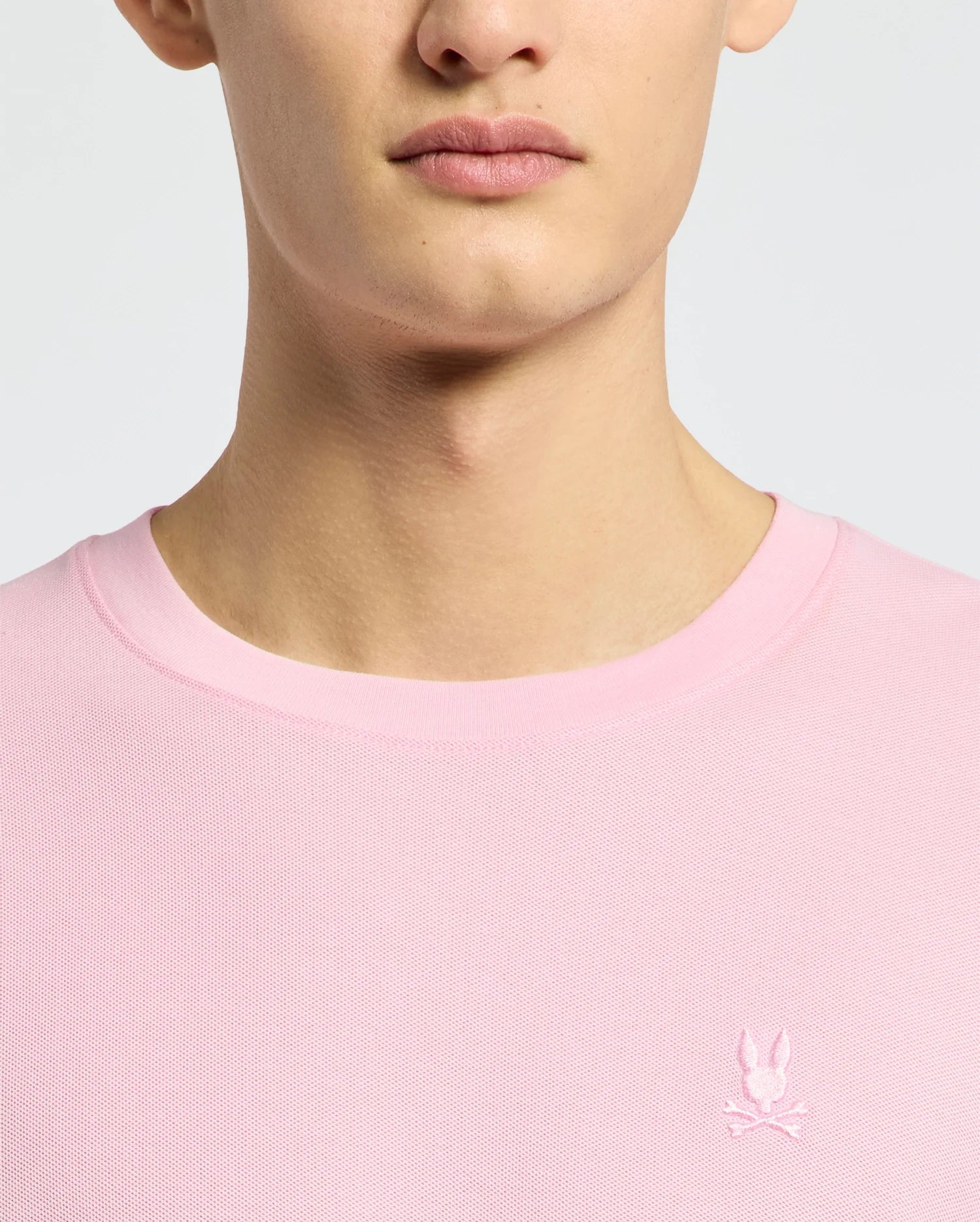 A person wears the Psycho Bunny MENS TAYLOR PIQUE TEE (B6U579F200), a light pink crew neck made from lightweight piqué, featuring a small white embroidered Bunny and crossed bones logo on the left chest. Only their lower face, neck, and upper torso are shown.