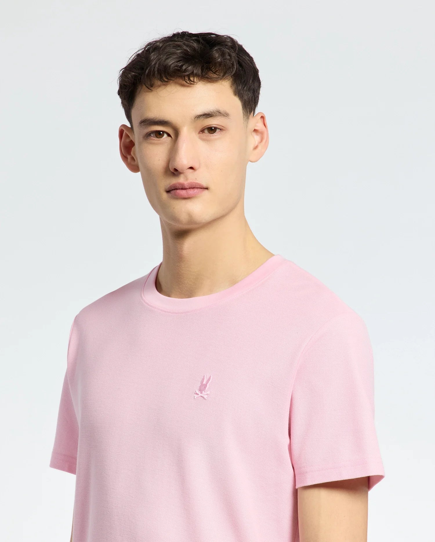 A young man with short brown hair wears the Psycho Bunny MENS TAYLOR PIQUE TEE (B6U579F200) in light pink piqué, featuring an embroidered chest Bunny, standing against a plain light background.