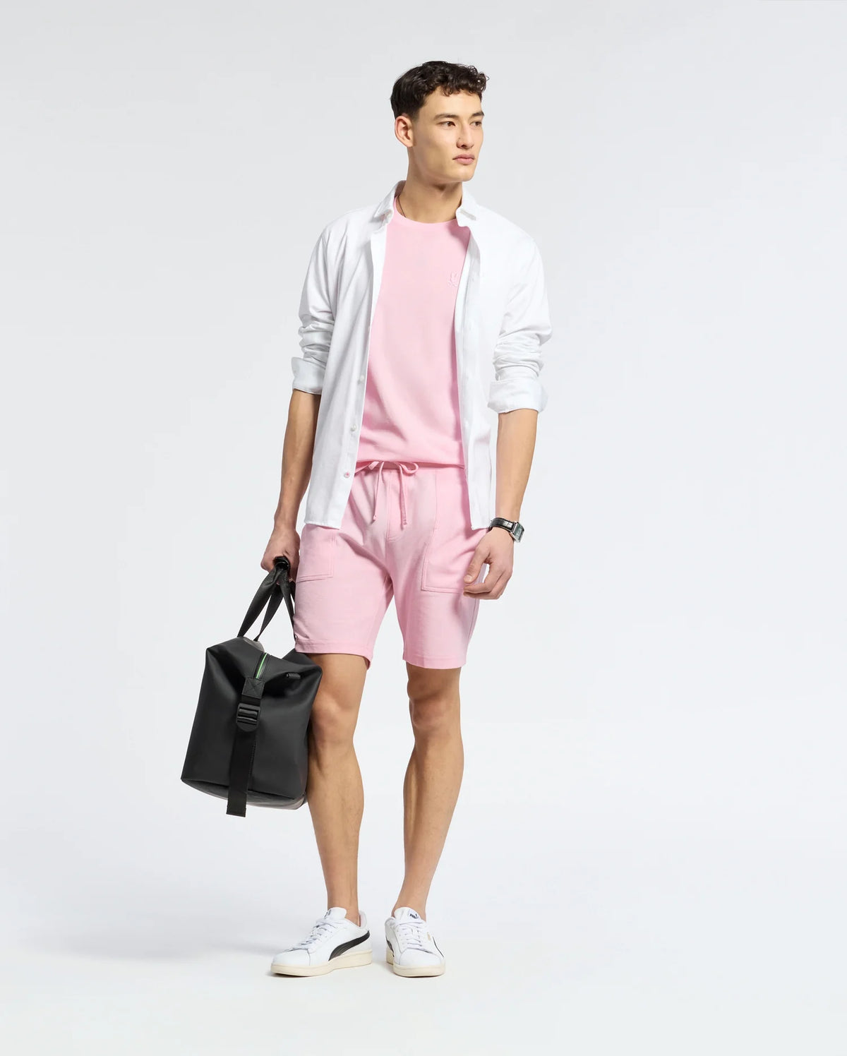 A young man stands against a plain background, wearing Psycho Bunny’s MENS TAYLOR PIQUE TEE (B6U579F200) in light pink with matching shorts, a white open button-down shirt with an embroidered Bunny, white sneakers, and holding a black duffel bag.