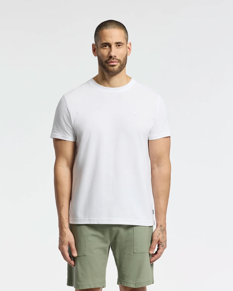 A short-haired, bearded individual stands against a white backdrop wearing a Psycho Bunny MENS TAYLOR PIQUE TEE in regular fit, crafted from lightweight Pima cotton, complemented by olive green shorts. Their relaxed stance is highlighted by the tee's embroidered chest bunny.