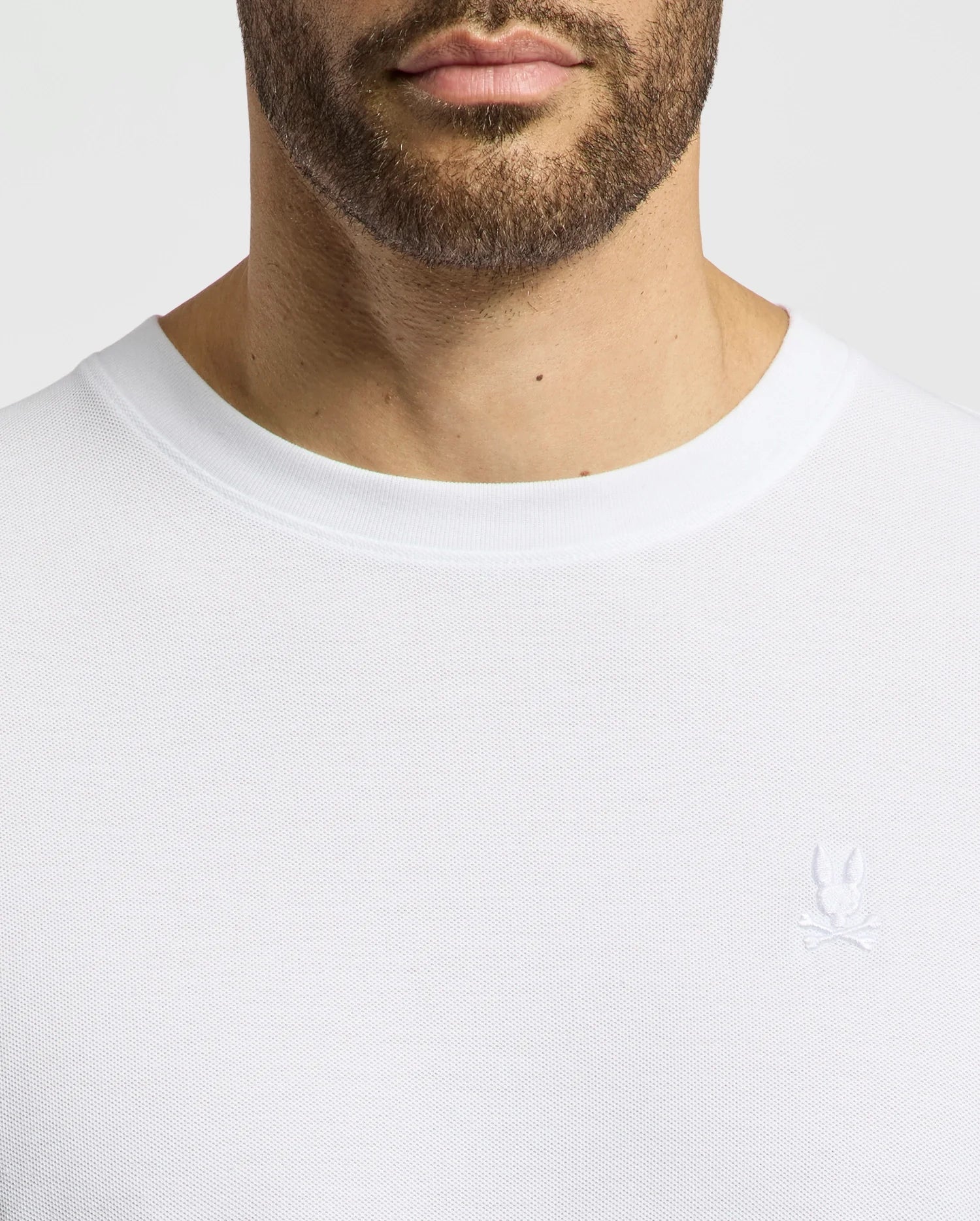 Close-up of a man wearing the Psycho Bunny MENS TAYLOR PIQUE TEE - B6U579F200, a regular-fit white crew-neck shirt in lightweight Pima cotton, featuring a small embroidered Bunny and crossbones emblem on the left chest, focusing on the upper torso and neckline.