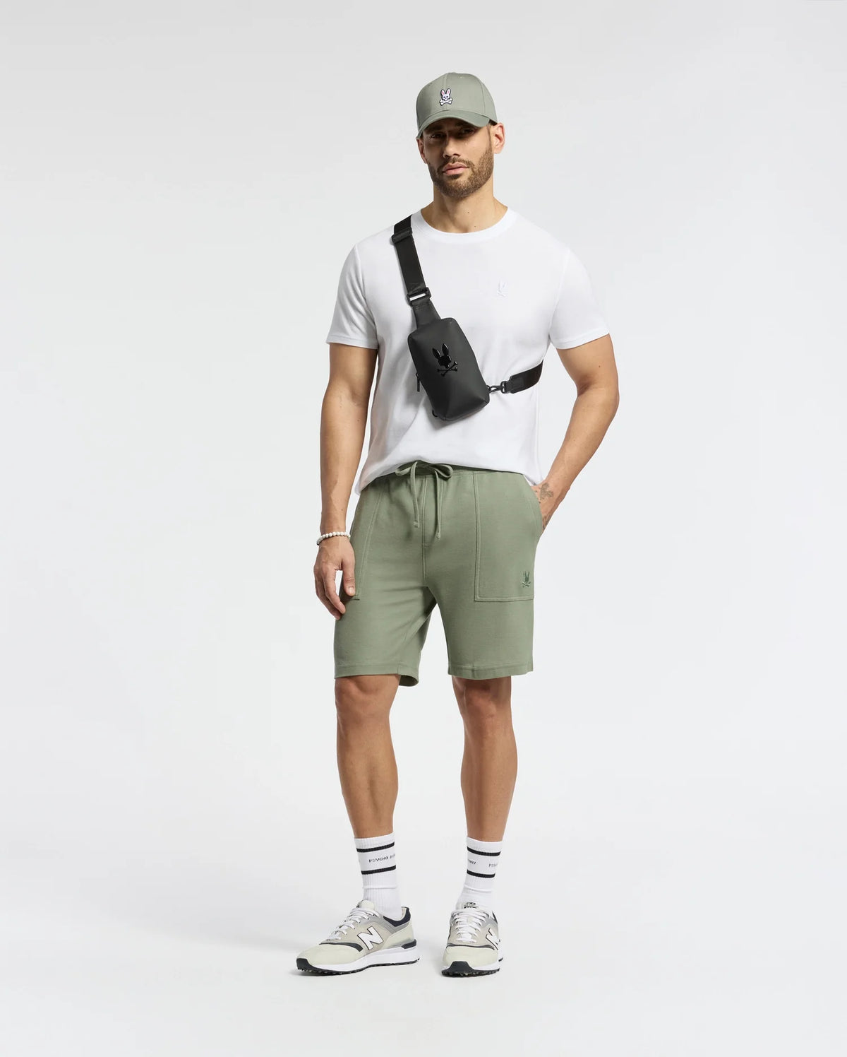 A man confidently stands against a plain backdrop, wearing the Psycho Bunny MENS TAYLOR PIQUE TEE in white with an embroidered bunny on the chest, a gray cap, sage green shorts, and gray sneakers. He accessorizes with a black crossbody bag and striped socks, hands in pockets.