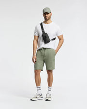 A man confidently stands against a plain backdrop, wearing the Psycho Bunny MENS TAYLOR PIQUE TEE in white with an embroidered bunny on the chest, a gray cap, sage green shorts, and gray sneakers. He accessorizes with a black crossbody bag and striped socks, hands in pockets.