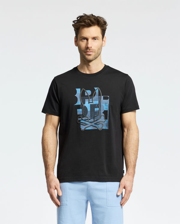 A man with short brown hair and a trimmed beard wears the Psycho Bunny MENS ZEALAND GRAPHIC TEE (B6U604F200), a black tee with a blue graphic, paired with light blue pants, standing against a plain white background.