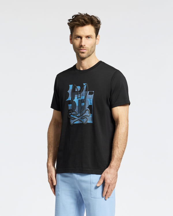 A man with short brown hair and a beard wears the Psycho Bunny MENS ZEALAND GRAPHIC TEE (B6U604F200) in black, paired with light blue pants, standing against a plain white background.