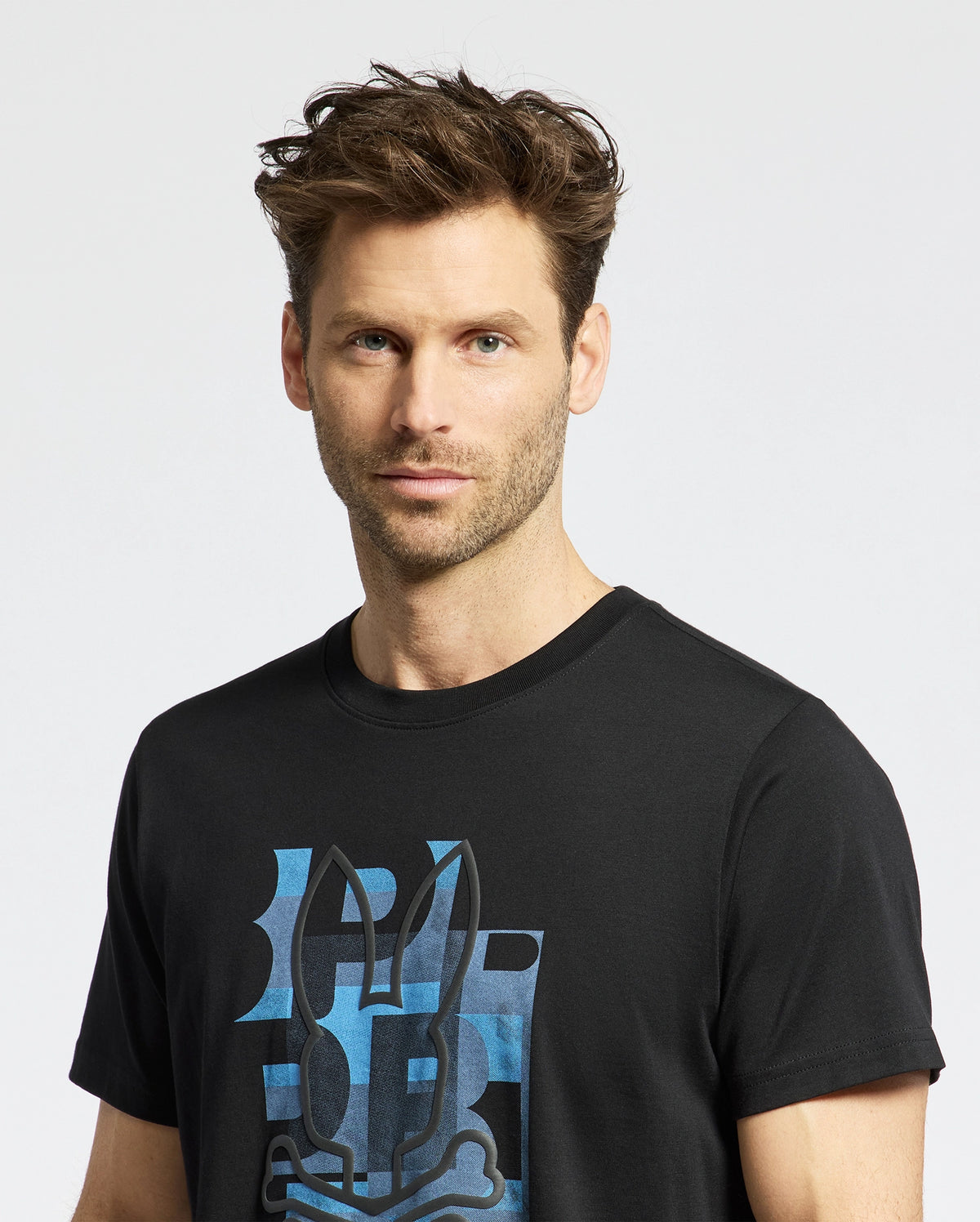 A man with tousled brown hair and light stubble wears the Psycho Bunny MENS ZEALAND GRAPHIC TEE—B6U604F200, a black shirt featuring a blue stylized bunny face. He stands against a white background, looking directly at the camera.