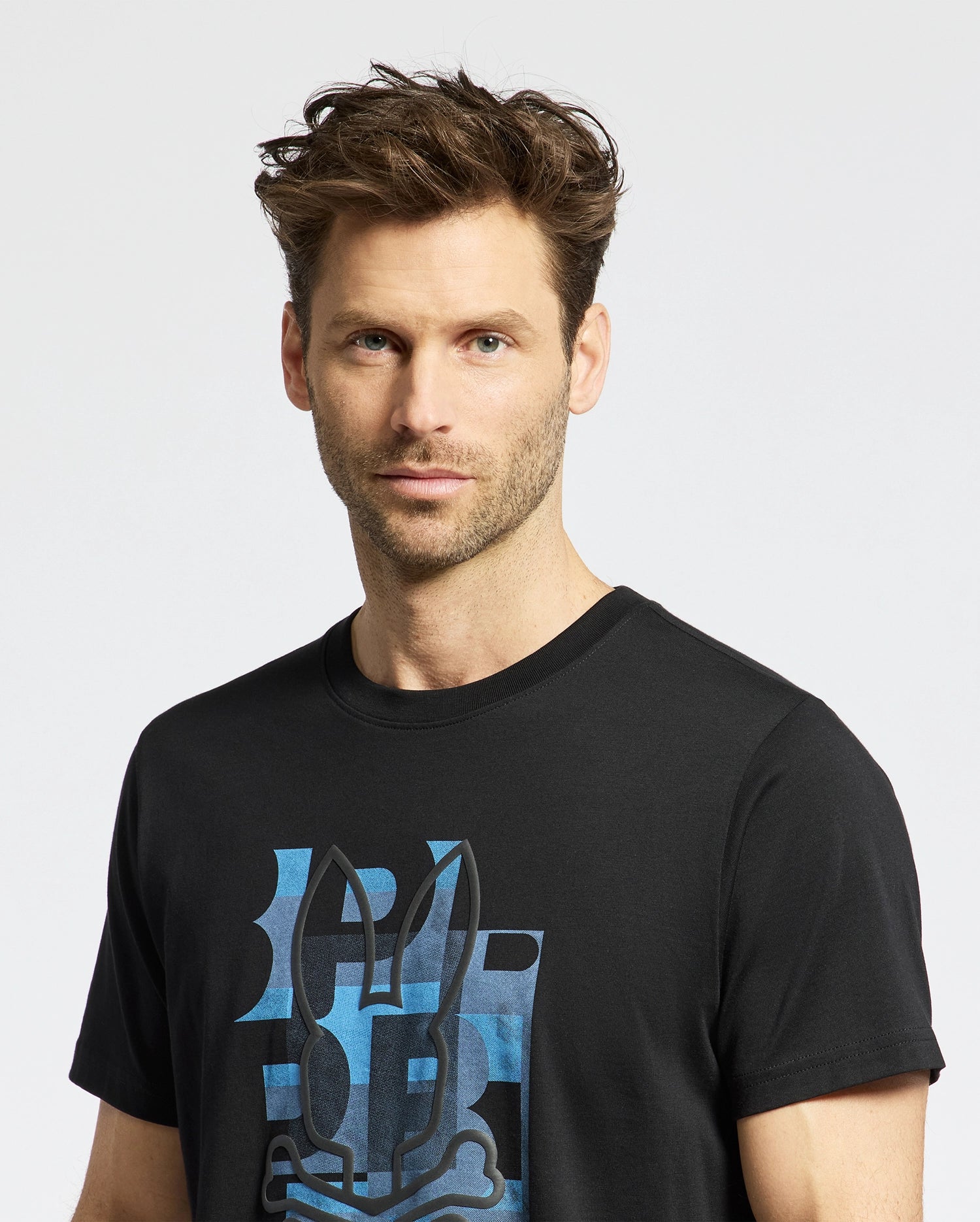 A man with tousled brown hair and light stubble wears the Psycho Bunny MENS ZEALAND GRAPHIC TEE—B6U604F200, a black shirt featuring a blue stylized bunny face. He stands against a white background, looking directly at the camera.