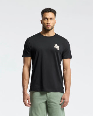 A man stands against a plain white background wearing Psycho Bunny's MENS ELLIOT FRONT BACK GRAPHIC TEE, featuring a small white water-based print design on the left chest, paired with light green pants.