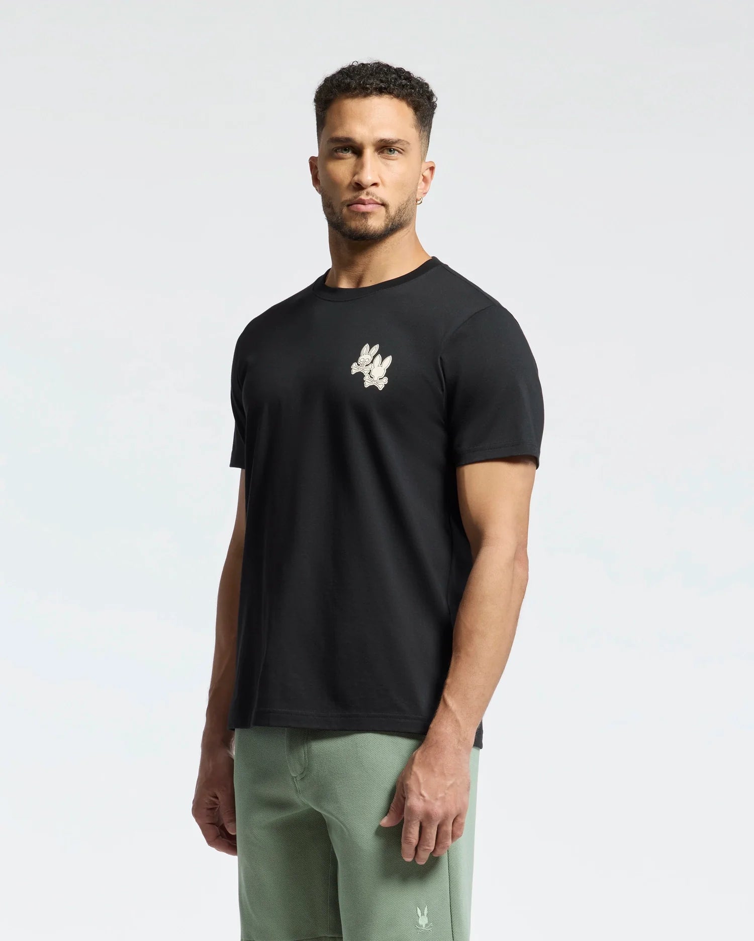 A man with short curly hair is wearing a Psycho Bunny MENS ELLIOT FRONT BACK GRAPHIC TEE in black, featuring a small white logo on the chest. Crafted from Pima cotton, it pairs perfectly with his light green shorts against a plain white background.
