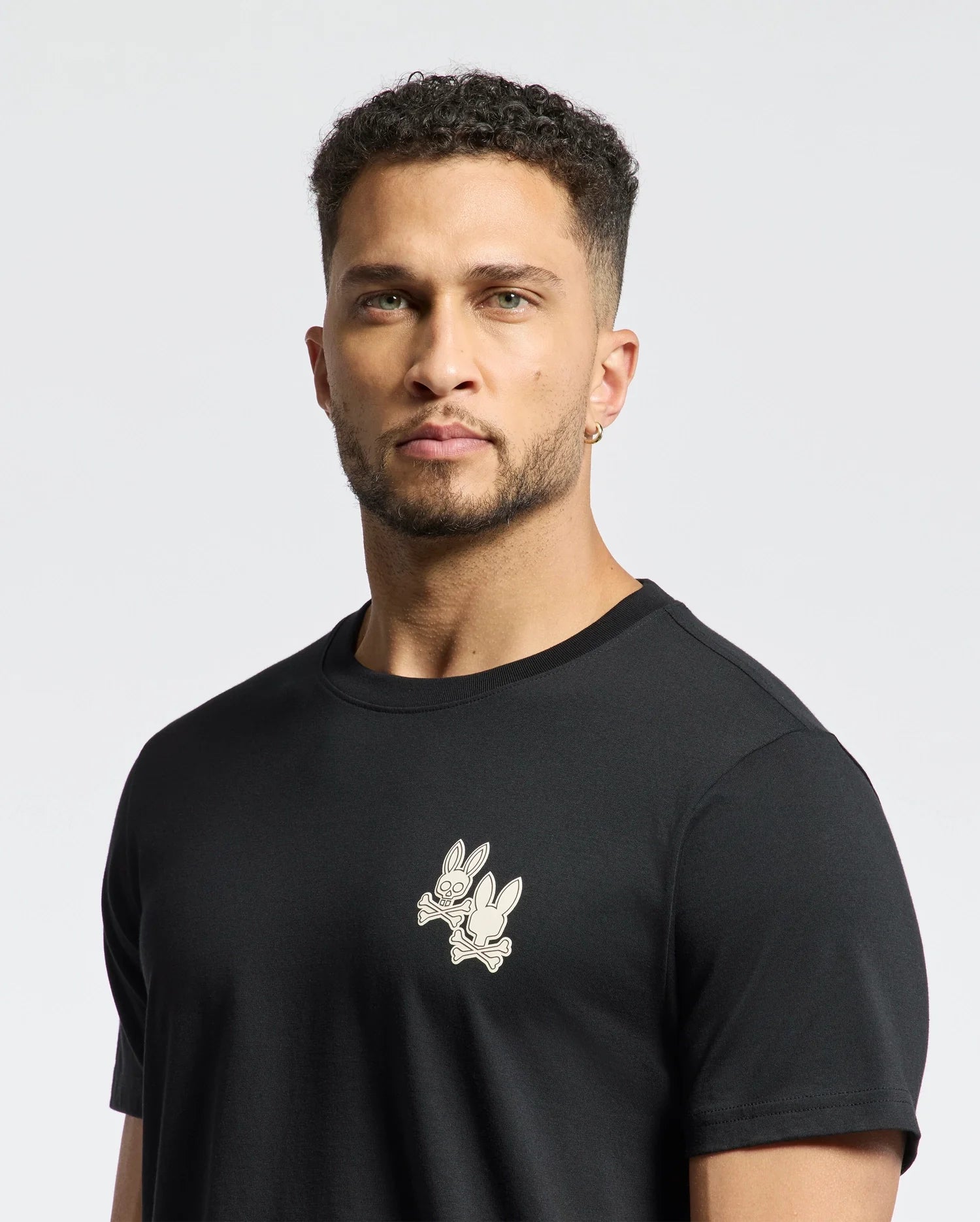 A man with curly hair and an earring models the Psycho Bunny Men's Elliot Front Back Graphic Tee (B6U605F200), a soft Pima cotton black T-shirt featuring a stylish rabbit print on the chest. He stands against a plain white background, looking directly at the camera.
