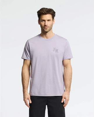 A man in Psycho Bunny's MENS ELLIOT FRONT BACK GRAPHIC TEE - B6U605F200, a light purple Pima cotton regular fit shirt with a small chest graphic, stands against a white background. He has short brown hair, a beard, and wears black pants.