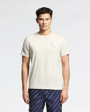 A man with short dark hair wears the Psycho Bunny MENS ELLIOT FRONT BACK GRAPHIC TEE - B6U605F200, a regular fit Pima cotton light beige t-shirt with a small white graphic print, and patterned dark blue shorts against a light gray background.