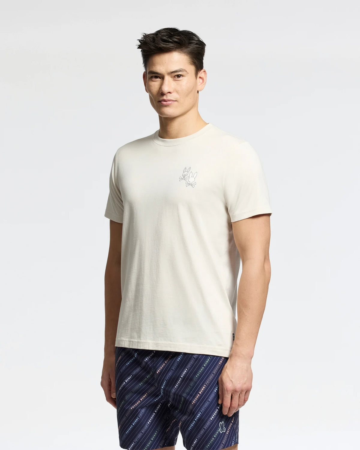 A man with short dark hair wears the Psycho Bunny MENS ELLIOT FRONT BACK GRAPHIC TEE - B6U605F200, a regular fit off-white Pima cotton tee with a small chest graphic, paired with patterned navy shorts against a light gray background.