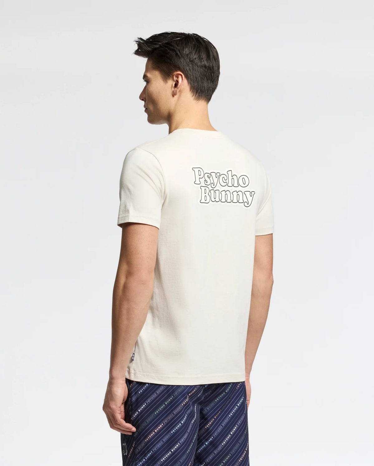 A man with short dark hair stands facing away, wearing the Psycho Bunny MENS ELLIOT FRONT BACK GRAPHIC TEE in cream and patterned navy shorts. The plain, light background highlights the tee's distinctive back graphic print.
