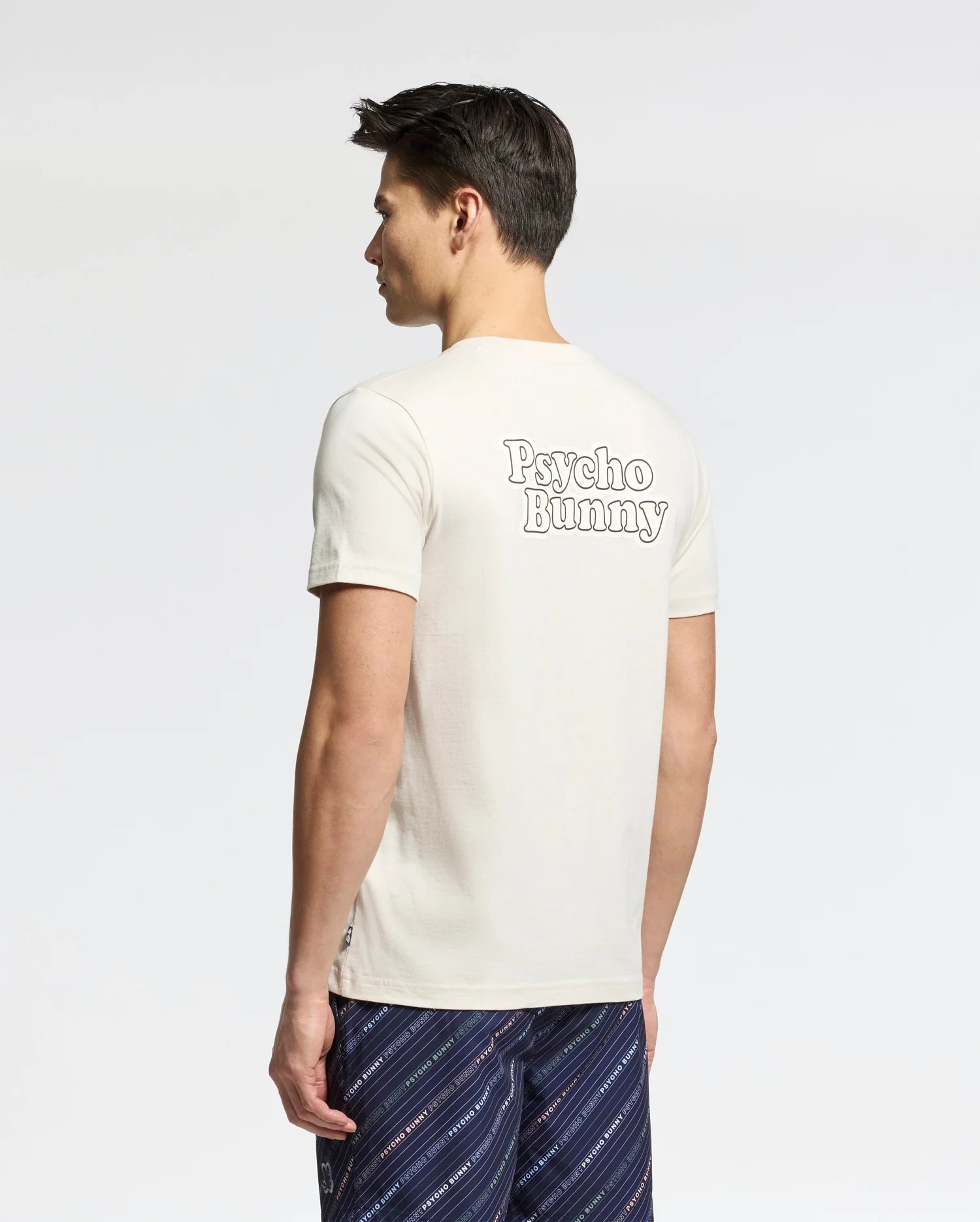 A man with short dark hair stands facing away, wearing the Psycho Bunny MENS ELLIOT FRONT BACK GRAPHIC TEE in cream and patterned navy shorts. The plain, light background highlights the tee's distinctive back graphic print.