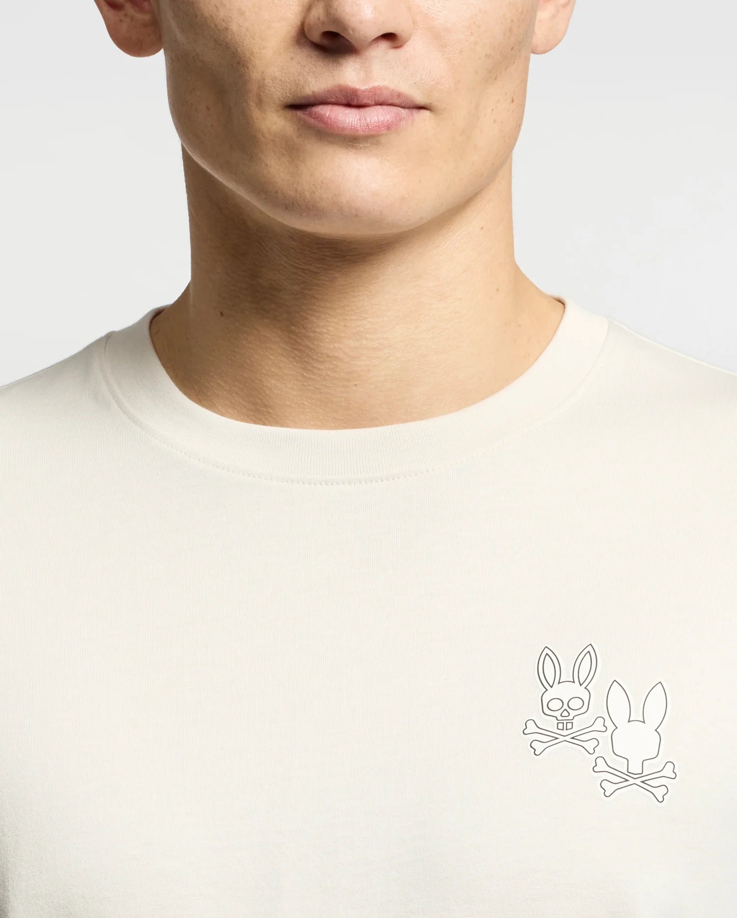 A person wearing the Psycho Bunny MENS ELLIOT FRONT BACK GRAPHIC TEE (B6U605F200), a regular fit, light-colored shirt featuring two bunny head and crossbones graphics—one on the left chest front, one on the back. Only face lower half and torso are shown.