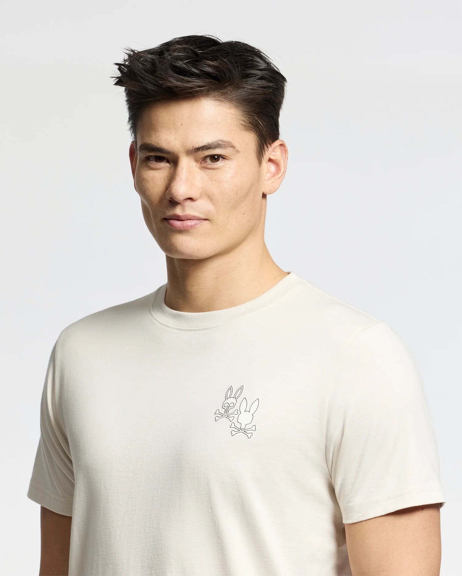 A man with short dark hair wears the Psycho Bunny MENS ELLIOT FRONT BACK GRAPHIC TEE (B6U605F200), a light Pima cotton shirt with a small outlined Bugs Bunny graphic on the chest, standing against a plain light background.