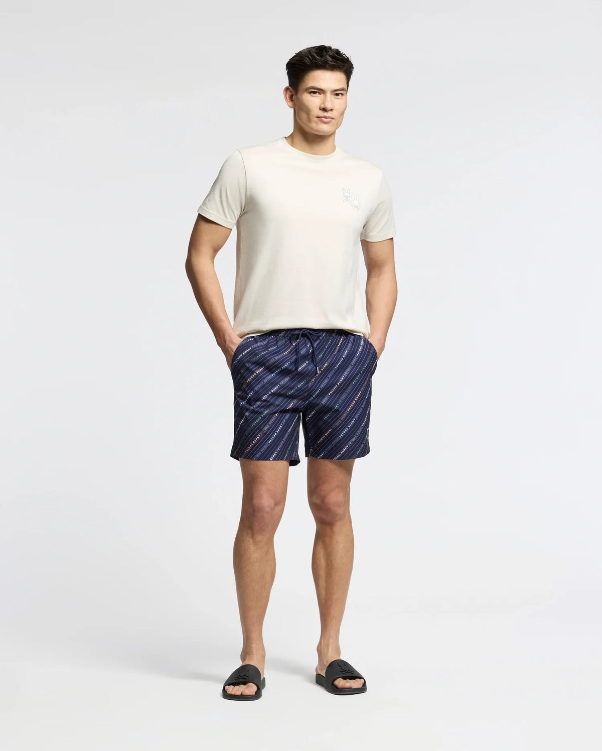 A man stands against a plain white background wearing the Psycho Bunny MENS ELLIOT FRONT BACK GRAPHIC TEE - B6U605F200, navy blue and white striped shorts, and black slide sandals, hands in pockets and looking relaxed.