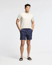 A man stands against a plain white background wearing the Psycho Bunny MENS ELLIOT FRONT BACK GRAPHIC TEE - B6U605F200, navy blue and white striped shorts, and black slide sandals, hands in pockets and looking relaxed.