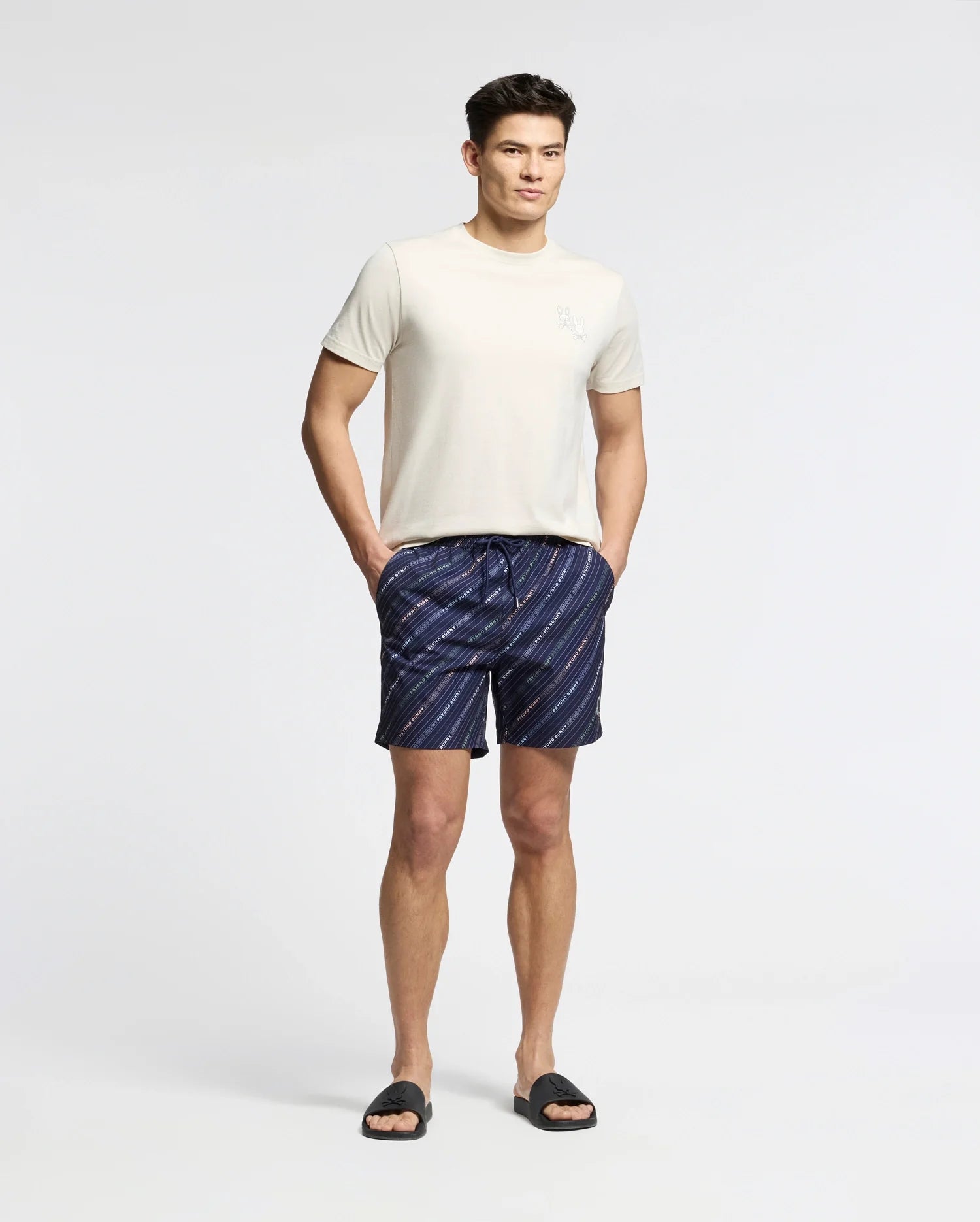 A man stands against a plain white background wearing the Psycho Bunny MENS ELLIOT FRONT BACK GRAPHIC TEE - B6U605F200, navy blue and white striped shorts, and black slide sandals, hands in pockets and looking relaxed.