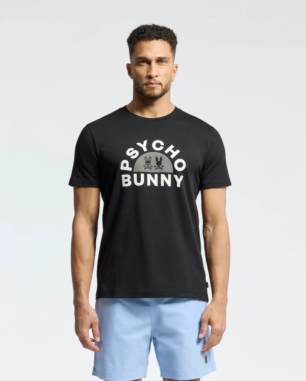 A man wears the Psycho Bunny MENS GOLDIE GRAPHIC TEE - B6U606F200 in black, featuring a bunny graphic, paired with light blue shorts against a plain white background.