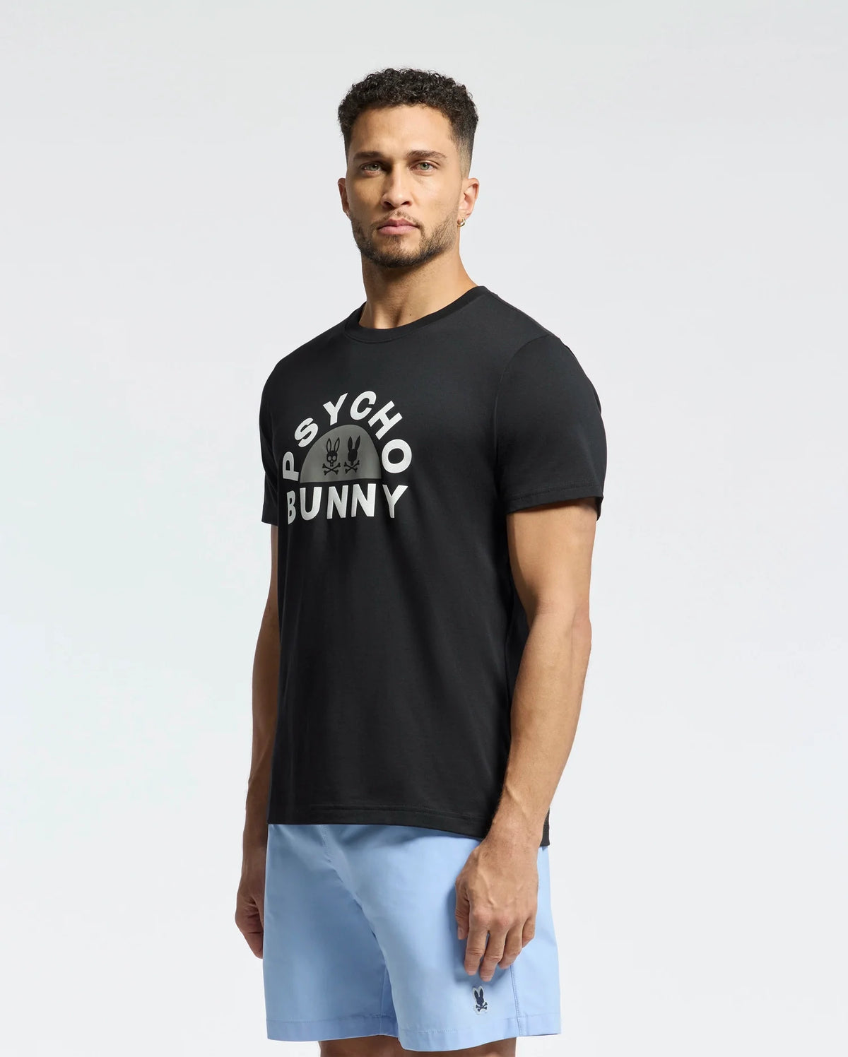 A man in a neutral pose wears the Psycho Bunny MENS GOLDIE GRAPHIC TEE - B6U606F200, crafted from premium Pima cotton, featuring white text and a subtle bunny graphic. He pairs it with light blue shorts against a plain background.