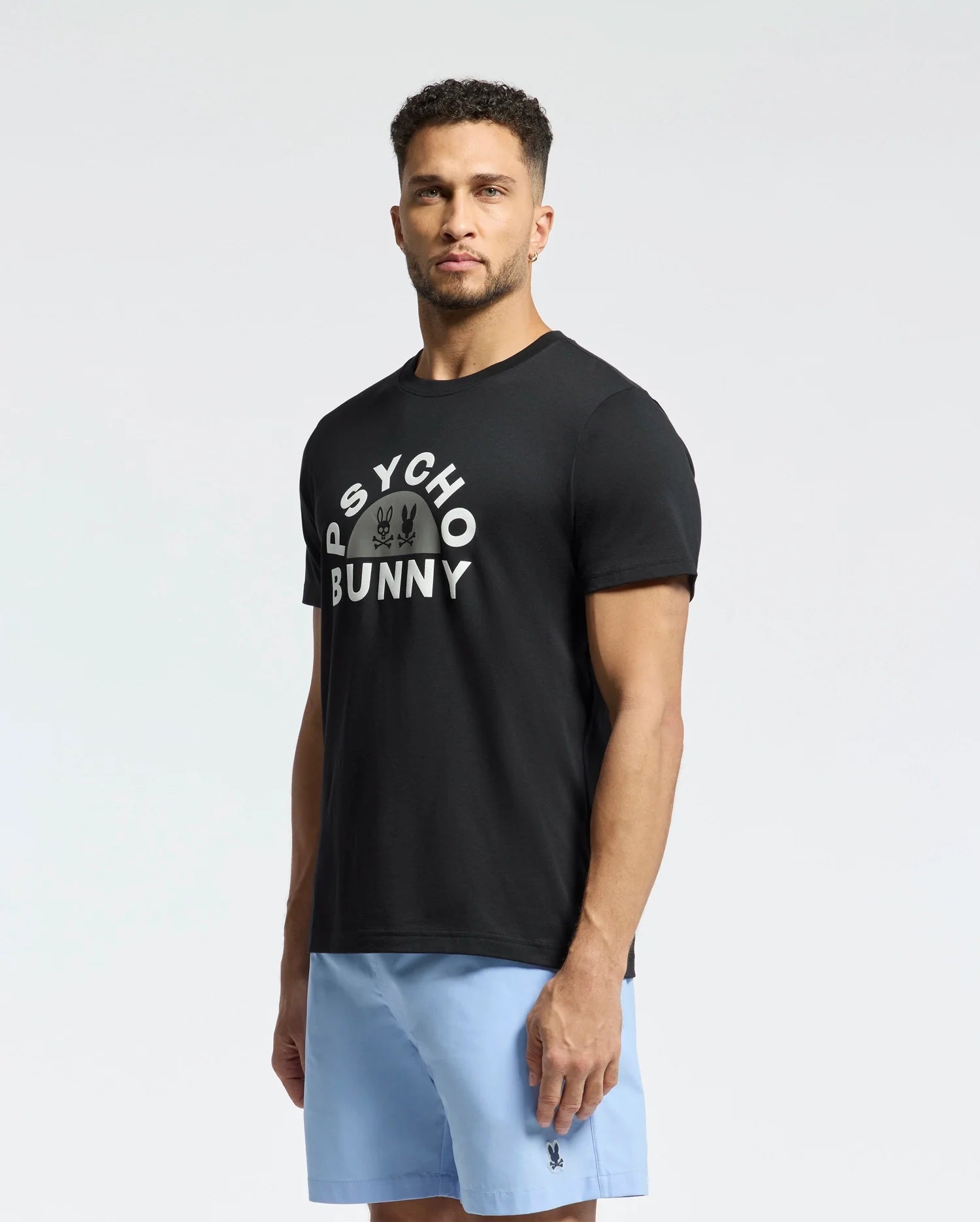 A man in a neutral pose wears the Psycho Bunny MENS GOLDIE GRAPHIC TEE - B6U606F200, crafted from premium Pima cotton, featuring white text and a subtle bunny graphic. He pairs it with light blue shorts against a plain background.