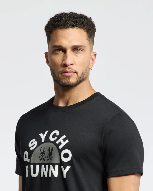 A man with short curly hair and an earring sports the MENS GOLDIE GRAPHIC TEE - B6U606F200 by Psycho Bunny, featuring "Psycho Bunny" text and a bold bunny graphic. The T-shirt, crafted from soft Pima cotton, pops against the plain white background.
