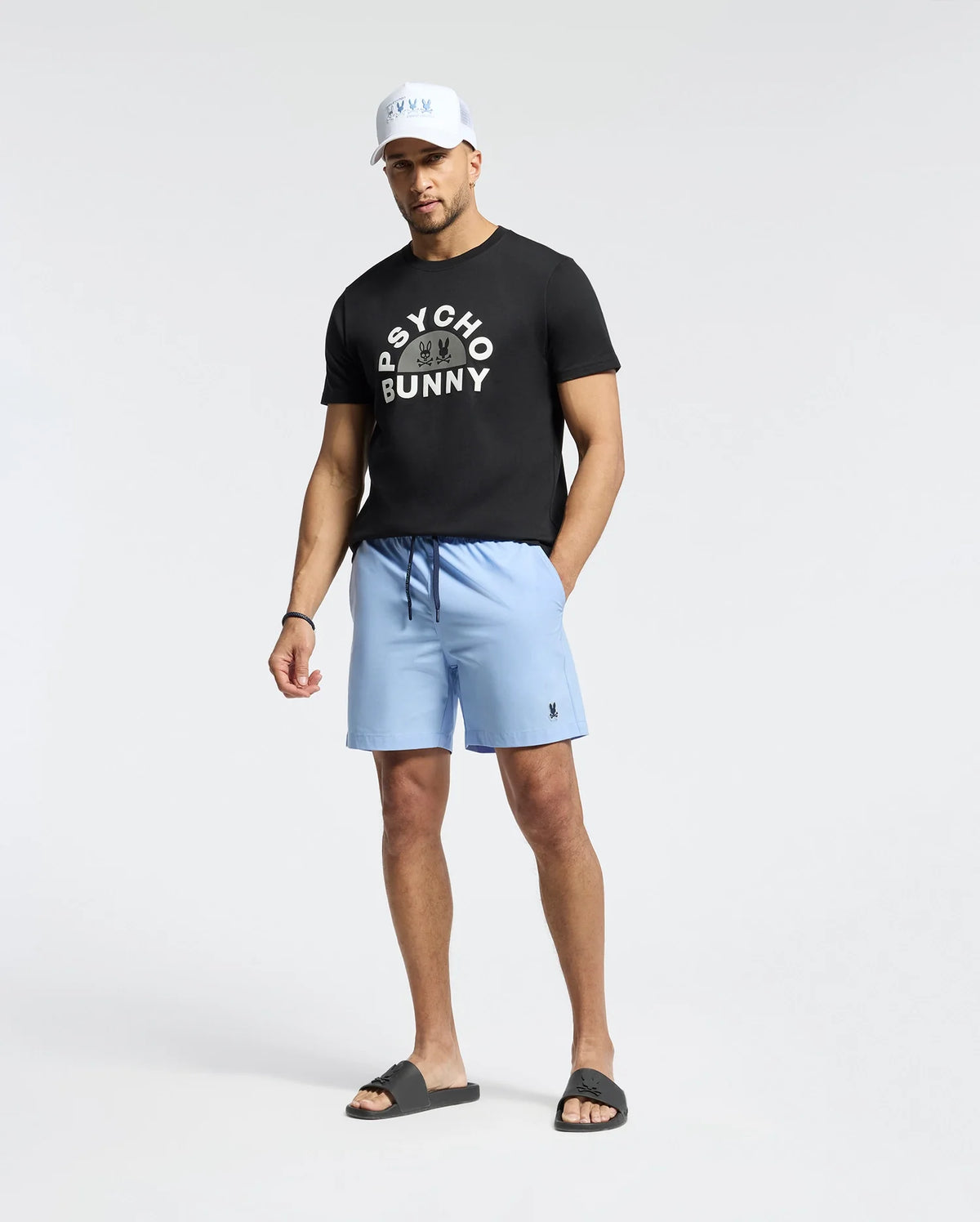 A man stands against a plain background wearing a Psycho Bunny MENS GOLDIE GRAPHIC TEE - B6U606F200, light blue shorts, black slides, and a white cap.