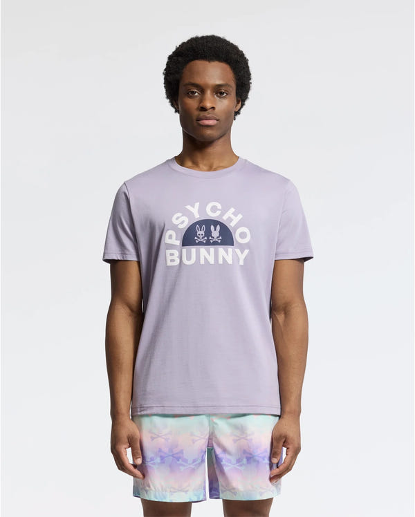 A man wears a light purple Psycho Bunny's Men's Goldie Graphic Tee (B6U606F200), adorned with a signature bunny design, paired with multicolored tie-dye shorts against a plain white background.