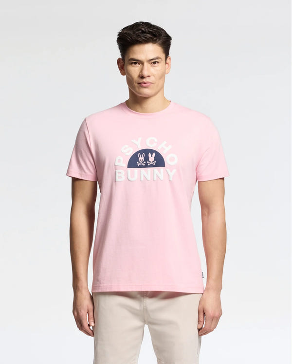 Wearing the Psycho Bunny MENS GOLDIE GRAPHIC TEE - B6U606F200, adorned with a vivid Bunny design and "Psycho Bunny" logo, a person stands out against the gray background. Crafted from soft Peruvian Pima cotton, this pink tee pairs perfectly with light-colored pants.