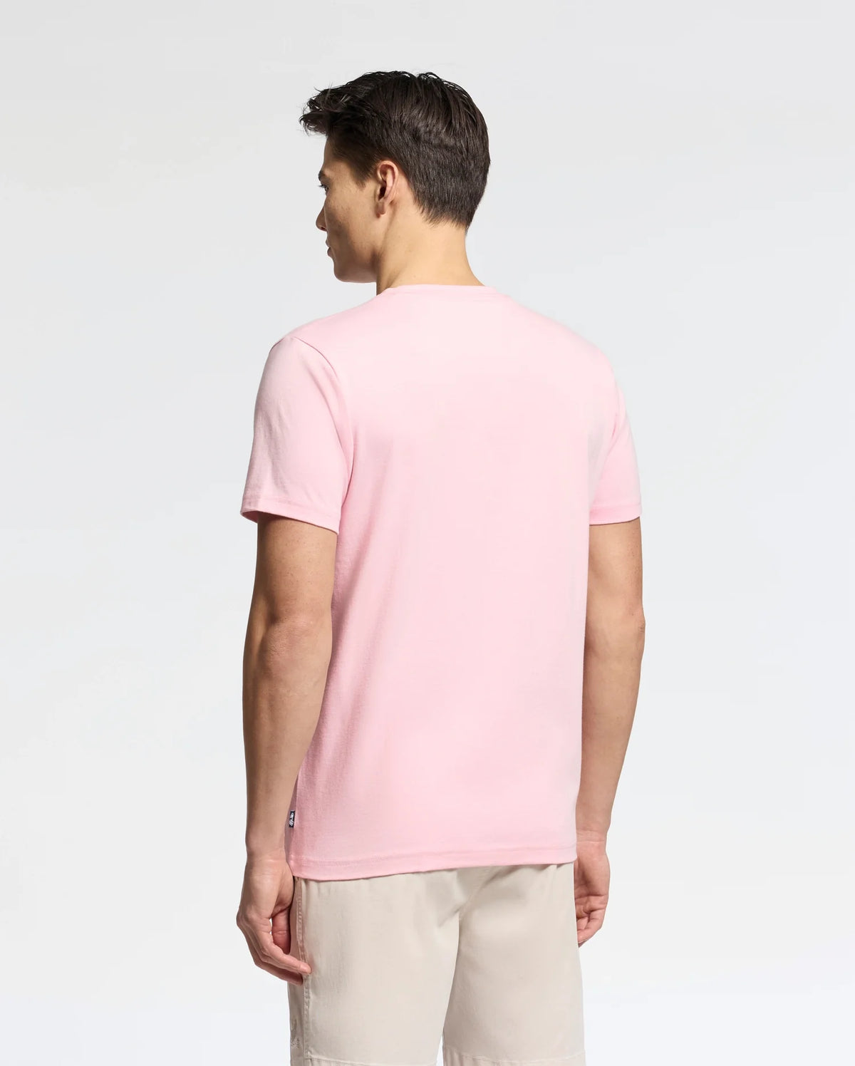A person with short dark hair wears a Psycho Bunny MENS GOLDIE GRAPHIC TEE - B6U606F200 made of soft Peruvian Pima cotton in light pink, paired with beige shorts, standing and facing away against a plain white background.