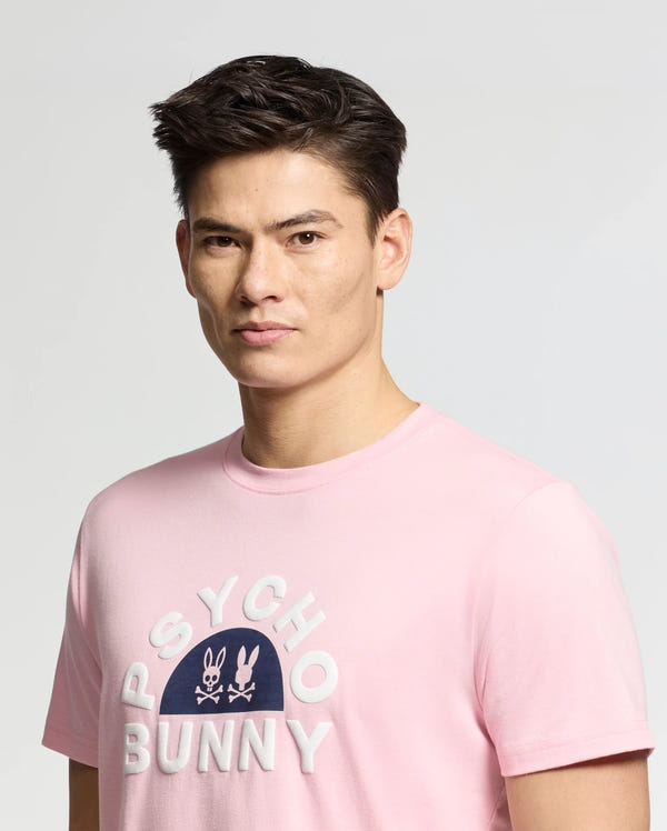 A person with short, dark hair is wearing a light pink MENS GOLDIE GRAPHIC TEE - B6U606F200 made of Peruvian Pima cotton, featuring "Psycho Bunny" and a bunny graphic on the front against a plain, light gray background.