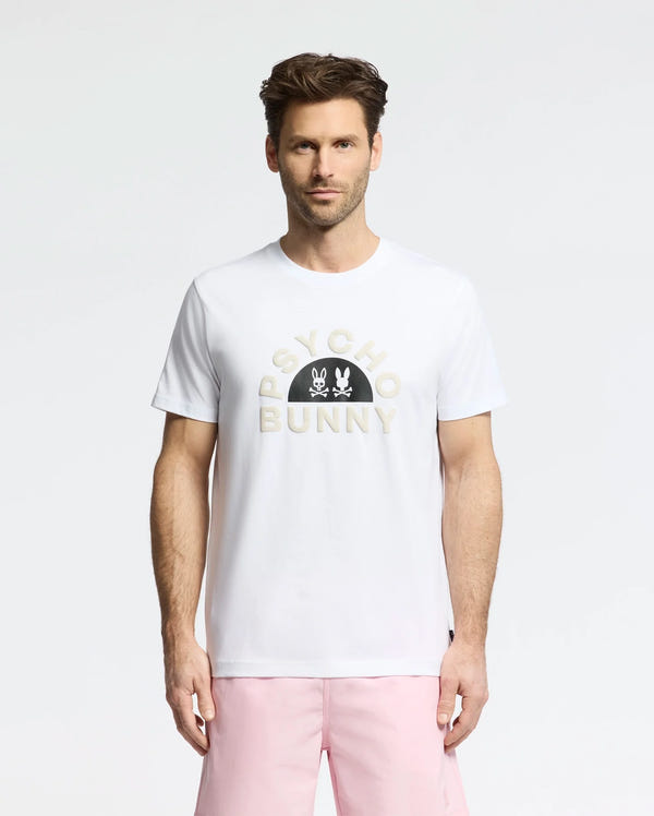 A person sports the "MENS GOLDIE GRAPHIC TEE - B6U606F200" by Psycho Bunny, a comfortable Peruvian cotton white t-shirt with a bold graphic, paired with light pink shorts.