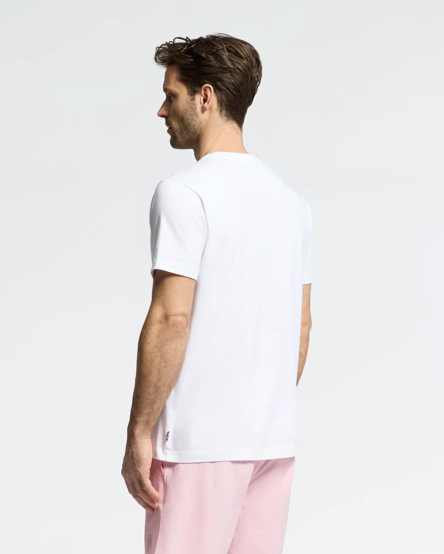 A man, with short brown hair, stands against a plain white background wearing the Psycho Bunny MENS GOLDIE GRAPHIC TEE (B6U606F200), a playful bunny on his white T-shirt, and pink shorts.