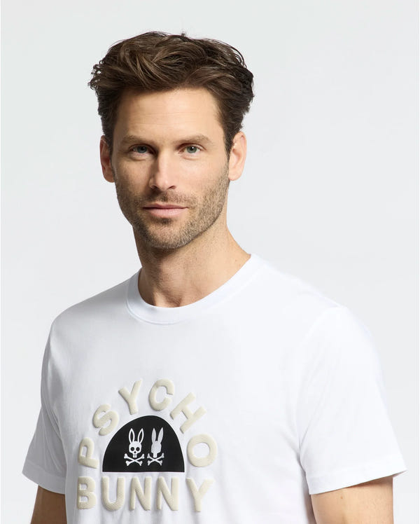 A man with short brown hair and a beard wears the MENS GOLDIE GRAPHIC TEE - B6U606F200 by Psycho Bunny, featuring a bunny graphic on a white Pima cotton shirt that highlights its unique design.