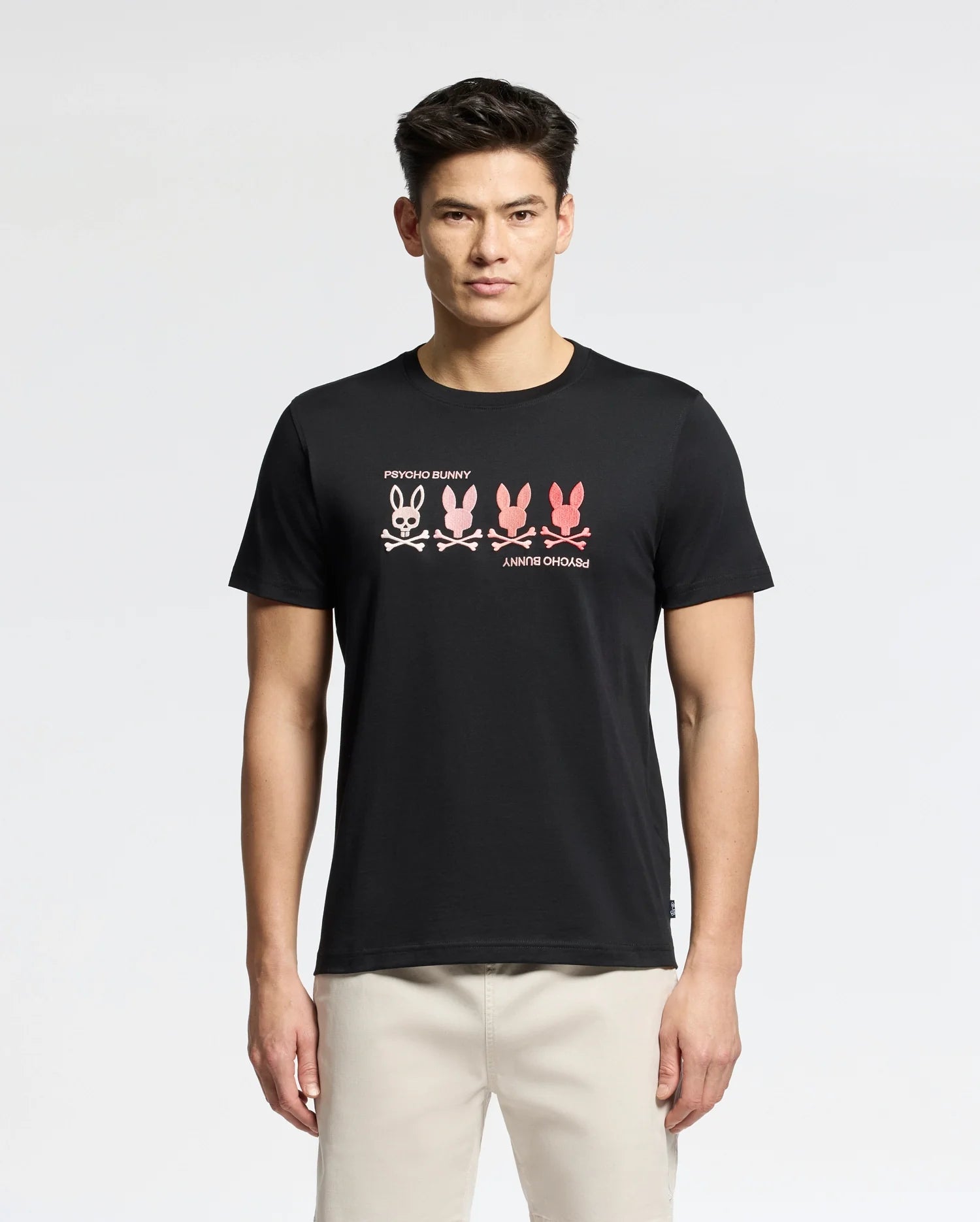 A man wears the Psycho Bunny MENS OCEANIA EMBROIDERED GRAPHIC TEE (B6U607F200) in black, showcasing a row of colorful abstract bunnies across the chest with retro vibes, paired with light pants.