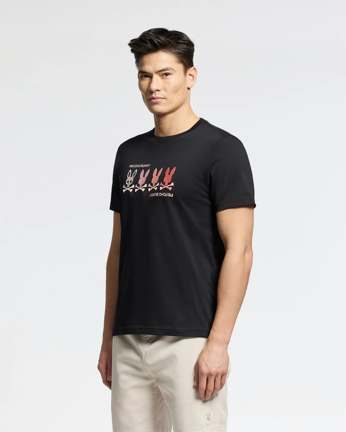 A man showcases a Psycho Bunny MENS OCEANIA EMBROIDERED GRAPHIC TEE in black, adorned with a vibrant row of multicolored bunny embroidery. He pairs it effortlessly with light trousers, and the white background accentuates the tee's subtle retro vibes.