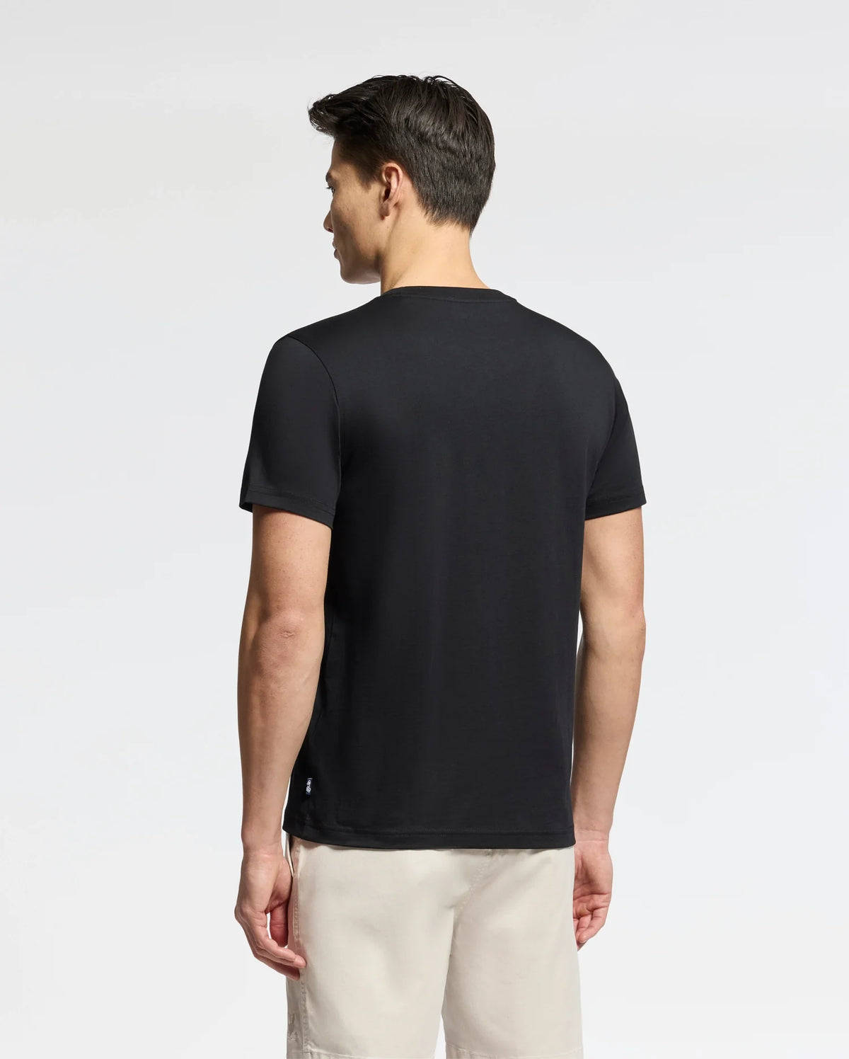 A man with short brown hair wears a Psycho Bunny Men's Oceania Embroidered Graphic Tee - B6U607F200 in black and beige shorts, standing against a neutral backdrop, evoking subtle retro vibes.
